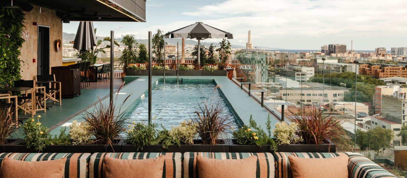 Rooftop pool at The Social Hub Barcelona Poblenou lined with striped orange and green cushions and umbrellas, overlooking the city skyline