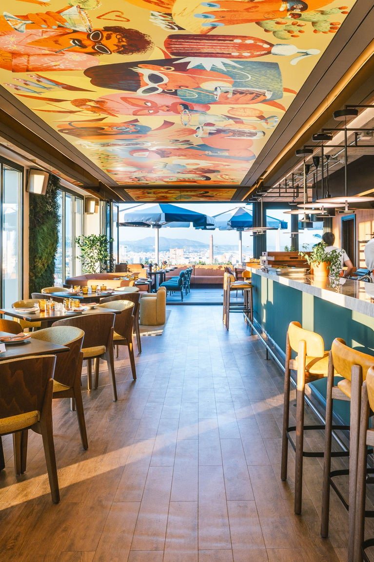 SISI rooftop restaurant at The Social Hub Barcelona Poblenou with a vivid illustrated mural ceiling, dining tables, and a bar counter overlooking the city