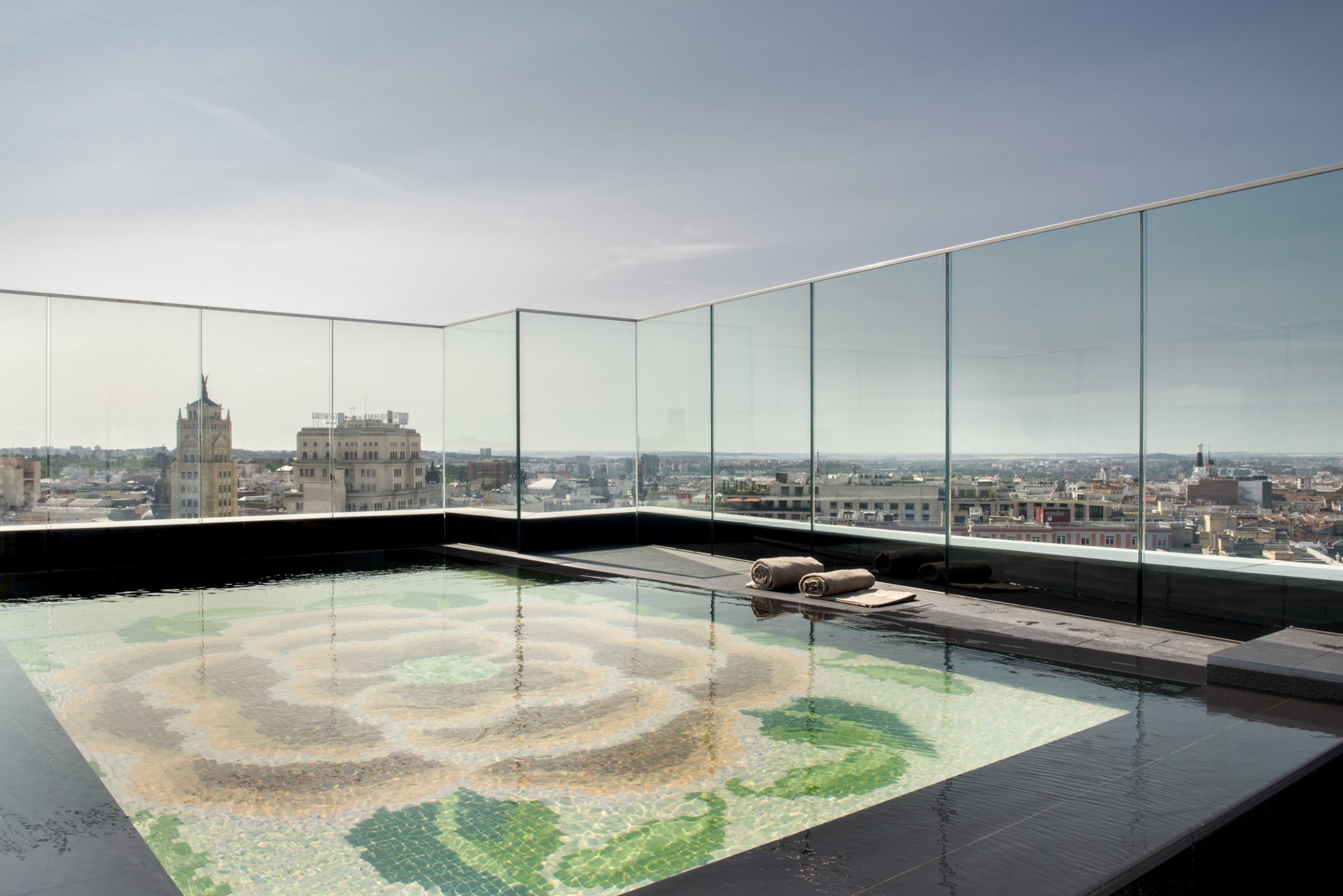 A mosaic-tiled plunge pool behind a glass balustrade, with the Madrid skyline beyond under a grey sky.