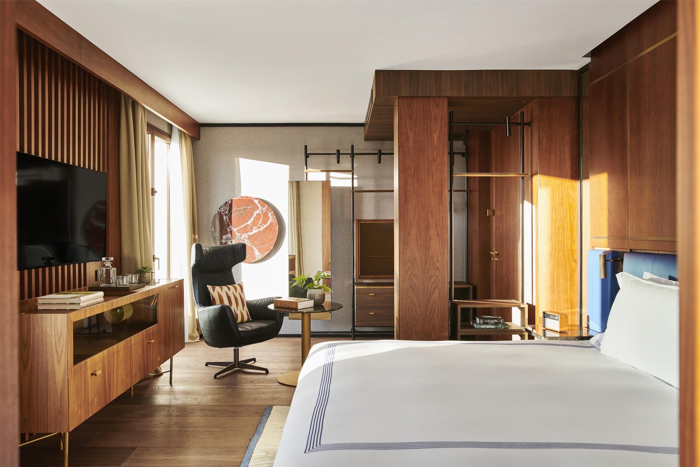 A guest room with walnut panelling, a white bed with a navy-striped throw, a black lounge chair and a wall-mounted TV.