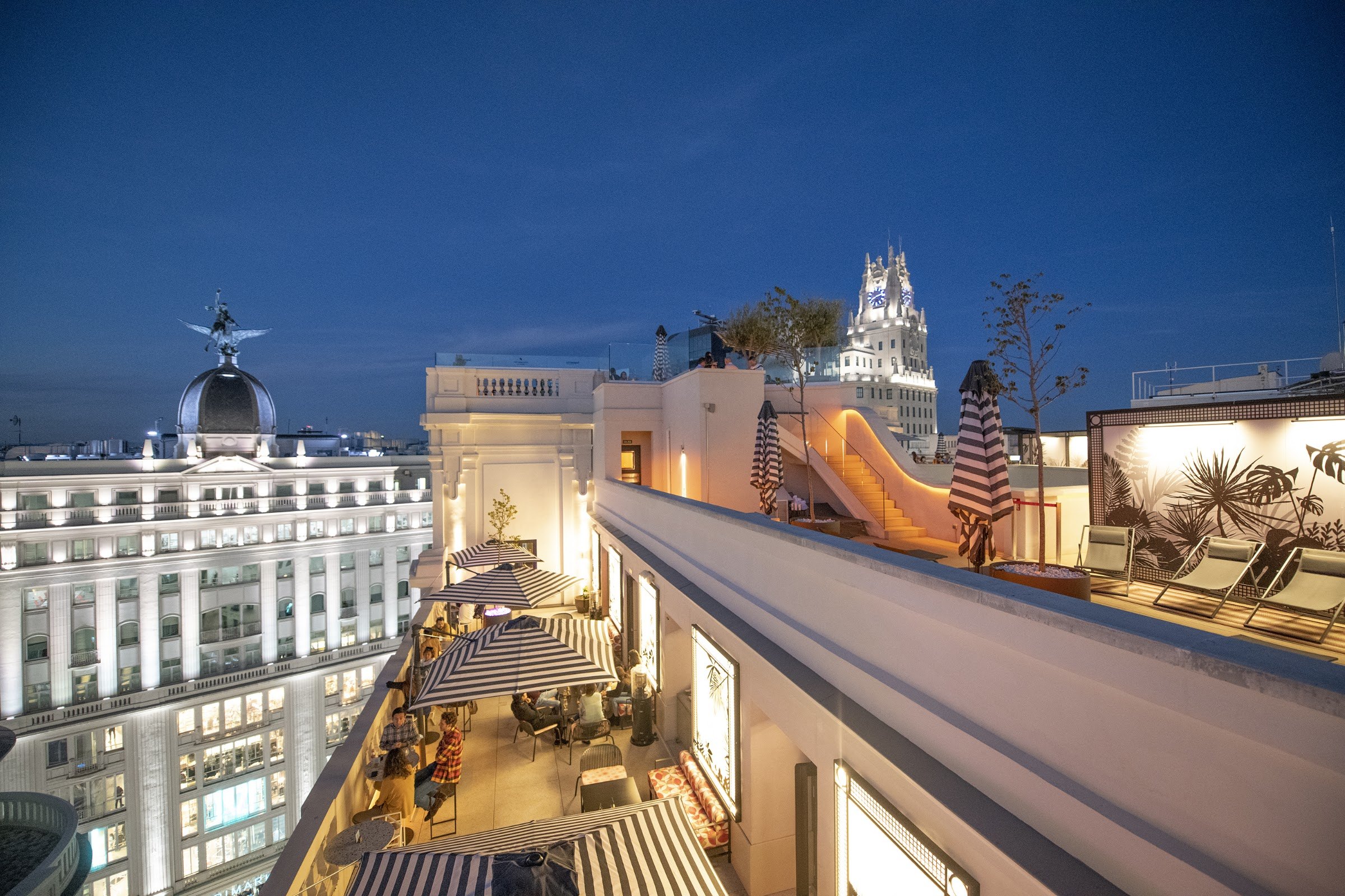 The rooftop terrace at dusk, with striped parasols and loungers looking over the lit rooftops and domes of central Madrid.