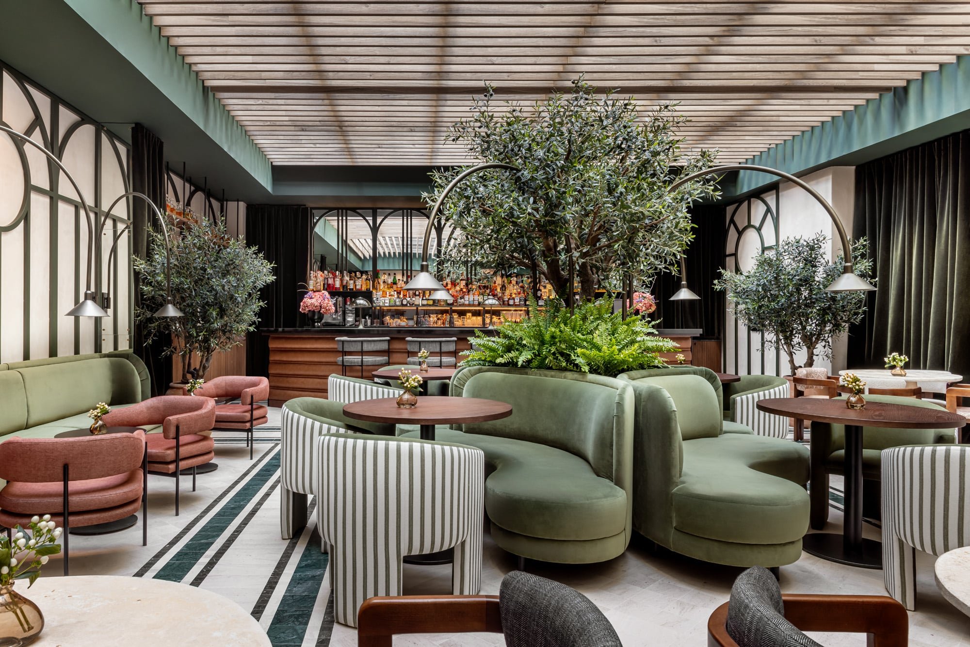 A lounge under a slatted wooden ceiling with olive trees, green velvet banquettes, striped armchairs and a bar with bottles in the background.