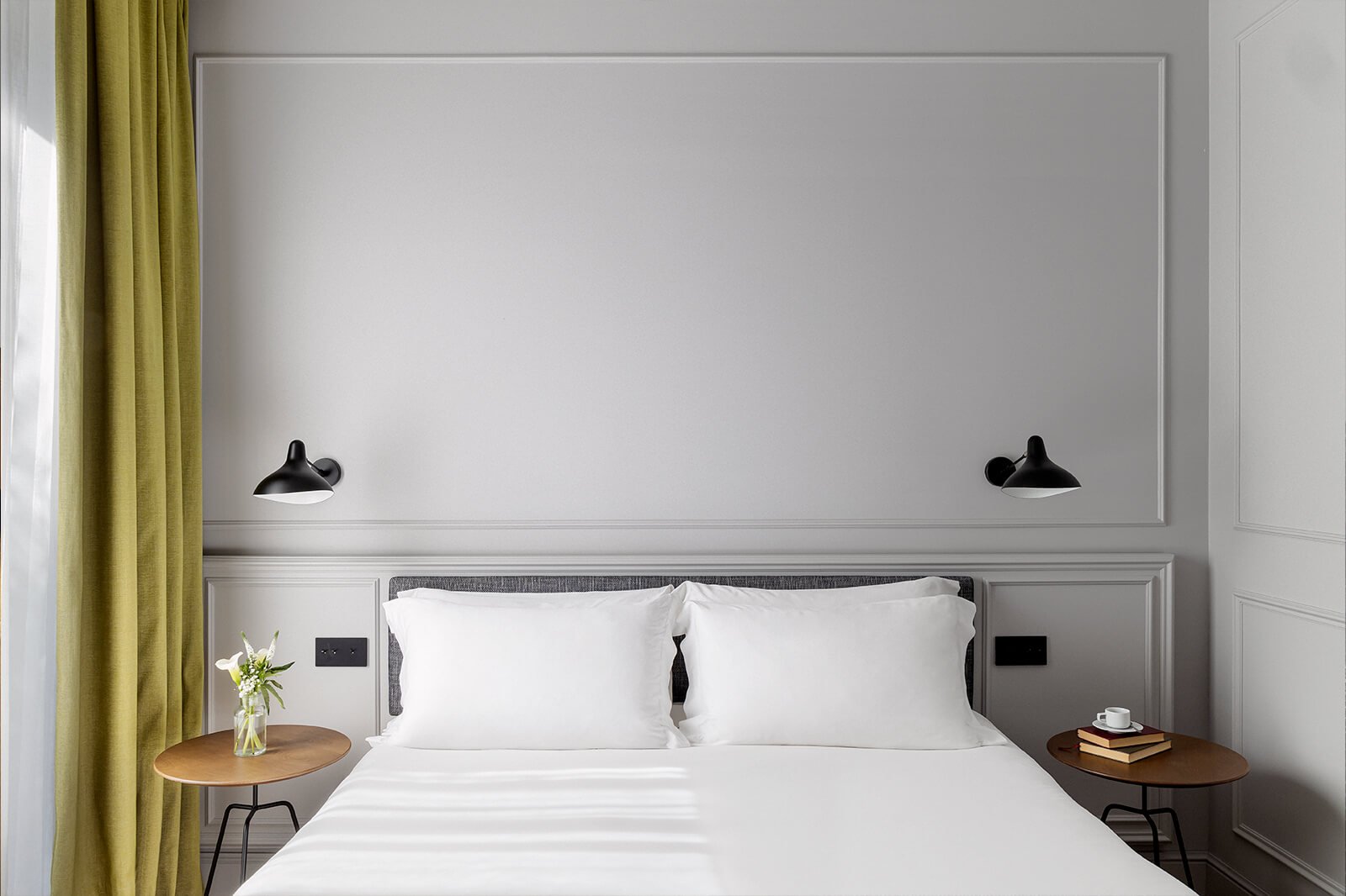 A white double bed with two wall lights on a grey panelled wall, a bedside table and an olive-green curtain.