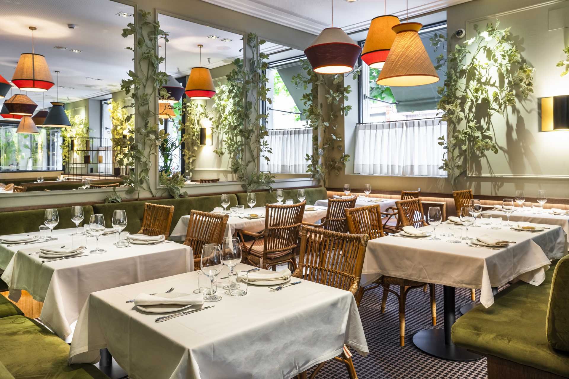 A restaurant dining room with tables set with white tablecloths and glassware, wicker chairs, hanging lamps and climbing plants along the windows.