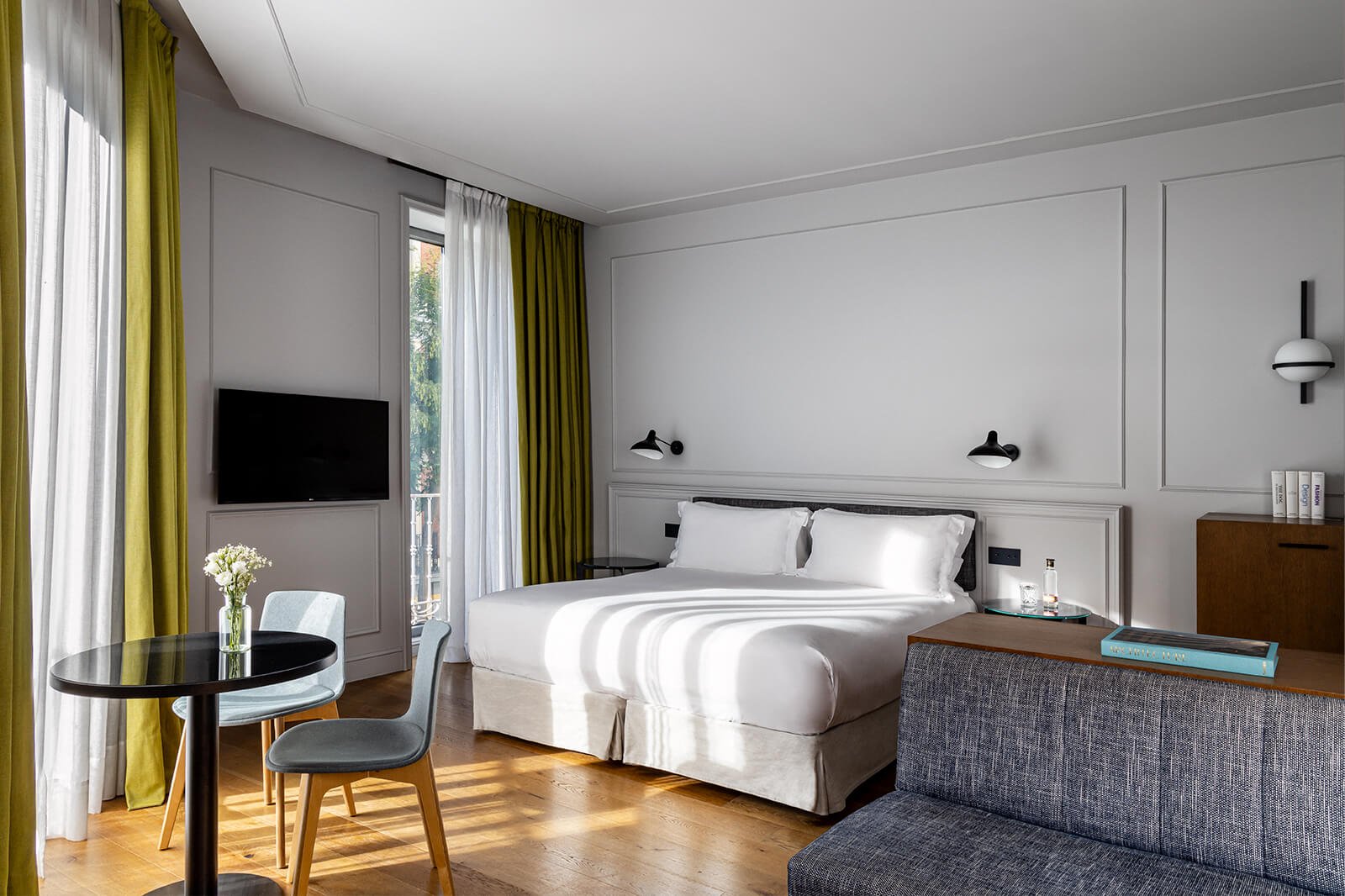 A hotel room with a large white bed, wall lights, a dark grey sofa, a small round table with two chairs and tall windows with olive-green curtains.