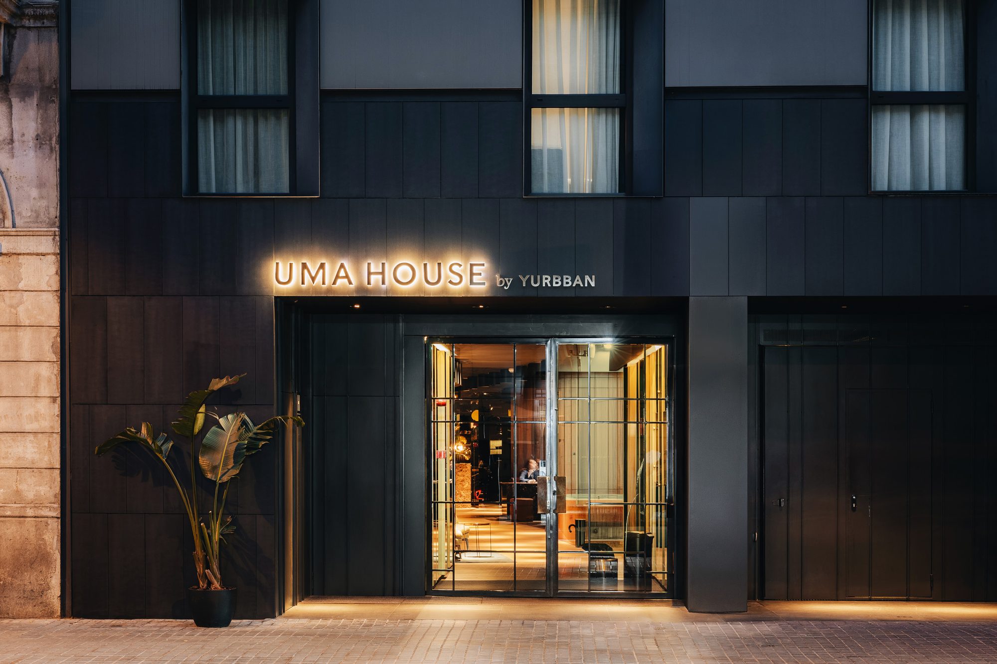 Dusk view of Uma House by Yurbban's dark charcoal facade with illuminated 'UMA HOUSE by YURBBAN' signage above the glass entrance doors