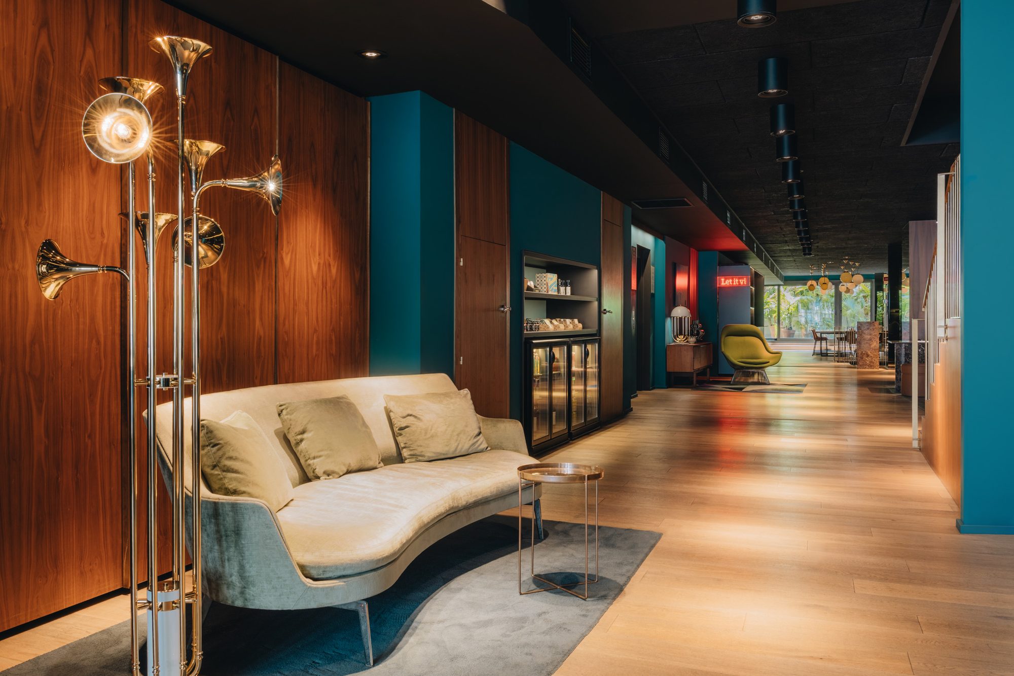 Lobby lounge at Uma House by Yurbban with a sculptural gold trumpet-shaped floor lamp, a curved cream velvet sofa and walnut wood paneling