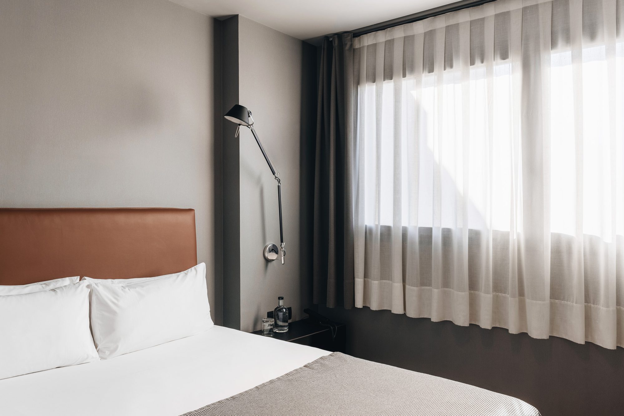 Guest room at Uma House by Yurbban with a cognac leather headboard, white bedding with a woven throw and an articulated black wall-mounted reading lamp