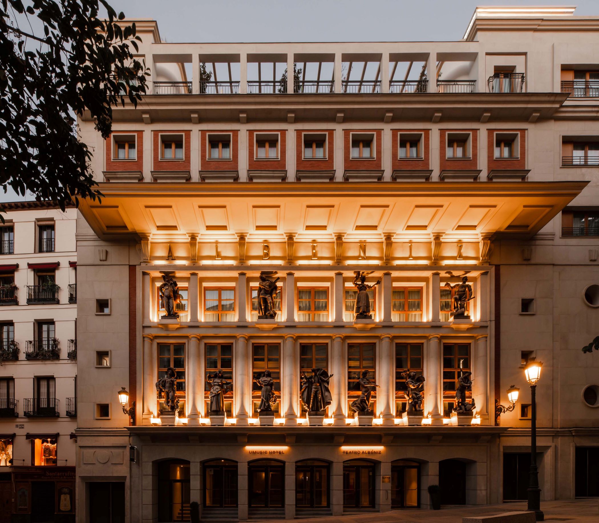 Illuminated stone facade at dusk with sculpted figures between columns, ALBENIZ lettering under the canopy, and entrance signage reading UMusic Hotel and Teatro Albeniz.
