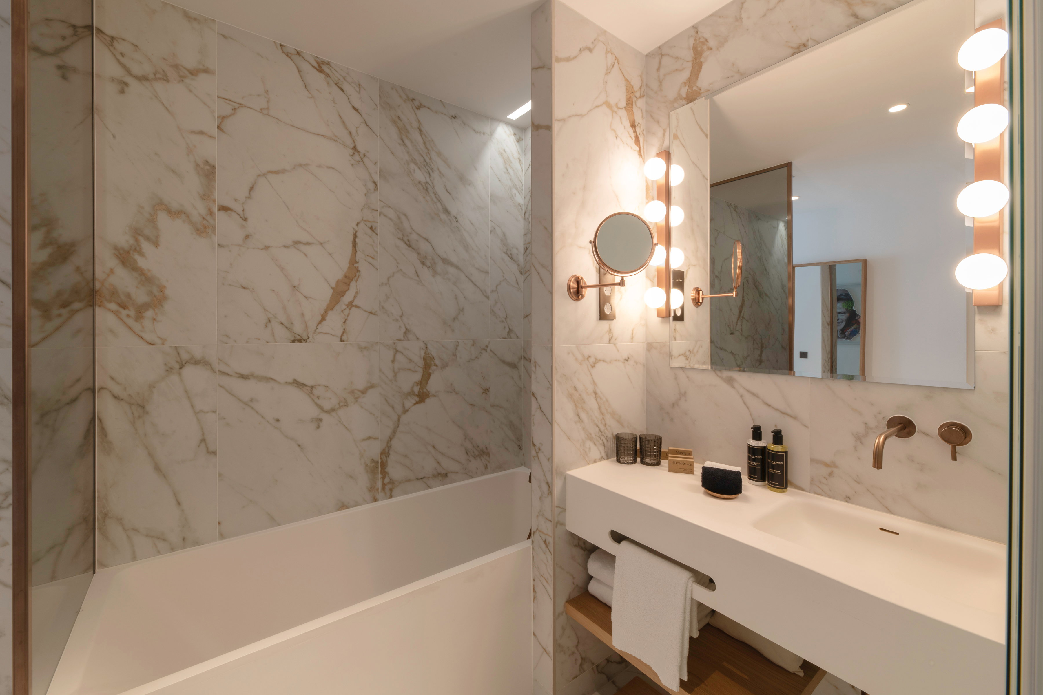 Marble guest bathroom with a bathtub, white stone vanity with toiletries and towels, round magnifying mirror, and globe-bulb vanity lights.