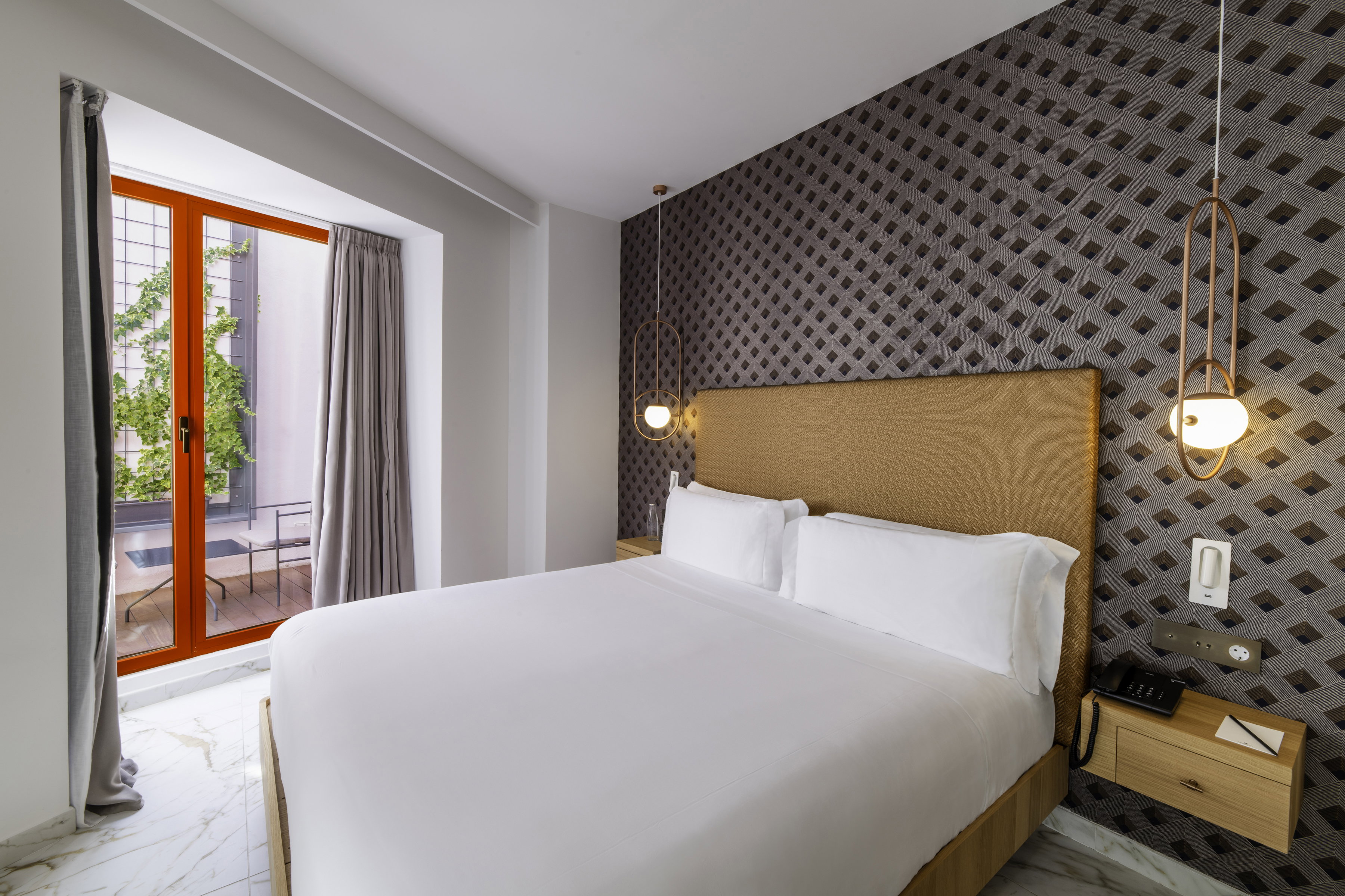 Guest room with a white-dressed double bed, gold fabric headboard, geometric-patterned feature wall, brass pendant lights, and an orange-framed glazed door onto a small balcony.