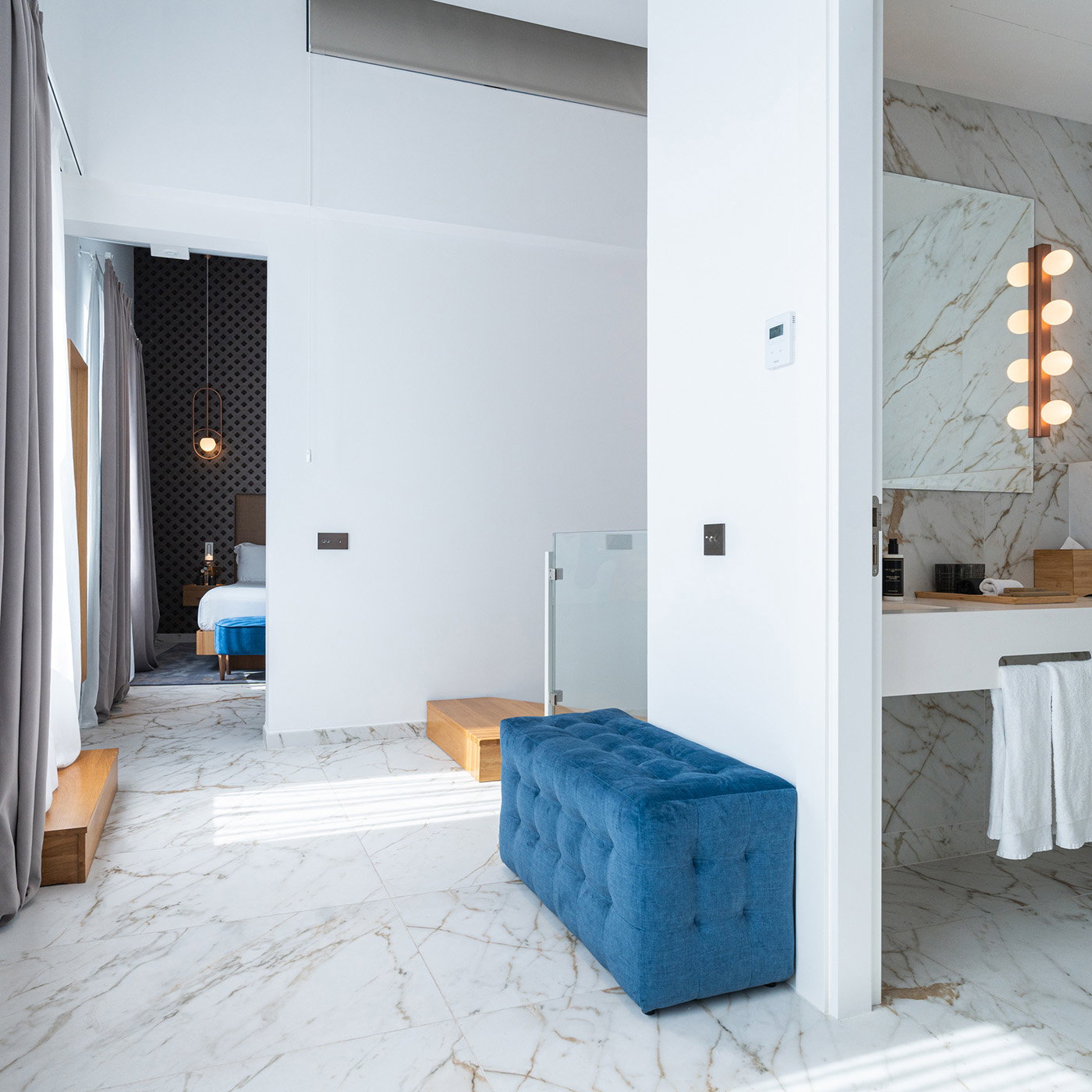 Marble-floored suite interior with a vanity counter, tufted grey ottoman, globe wall lights, and a bed visible in the adjoining room.