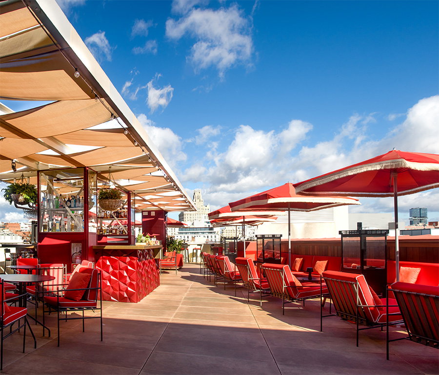 Rooftop terrace with red banquette seating, black metal chairs, fringed parasols and shade sails, and Madrid rooftops beyond.