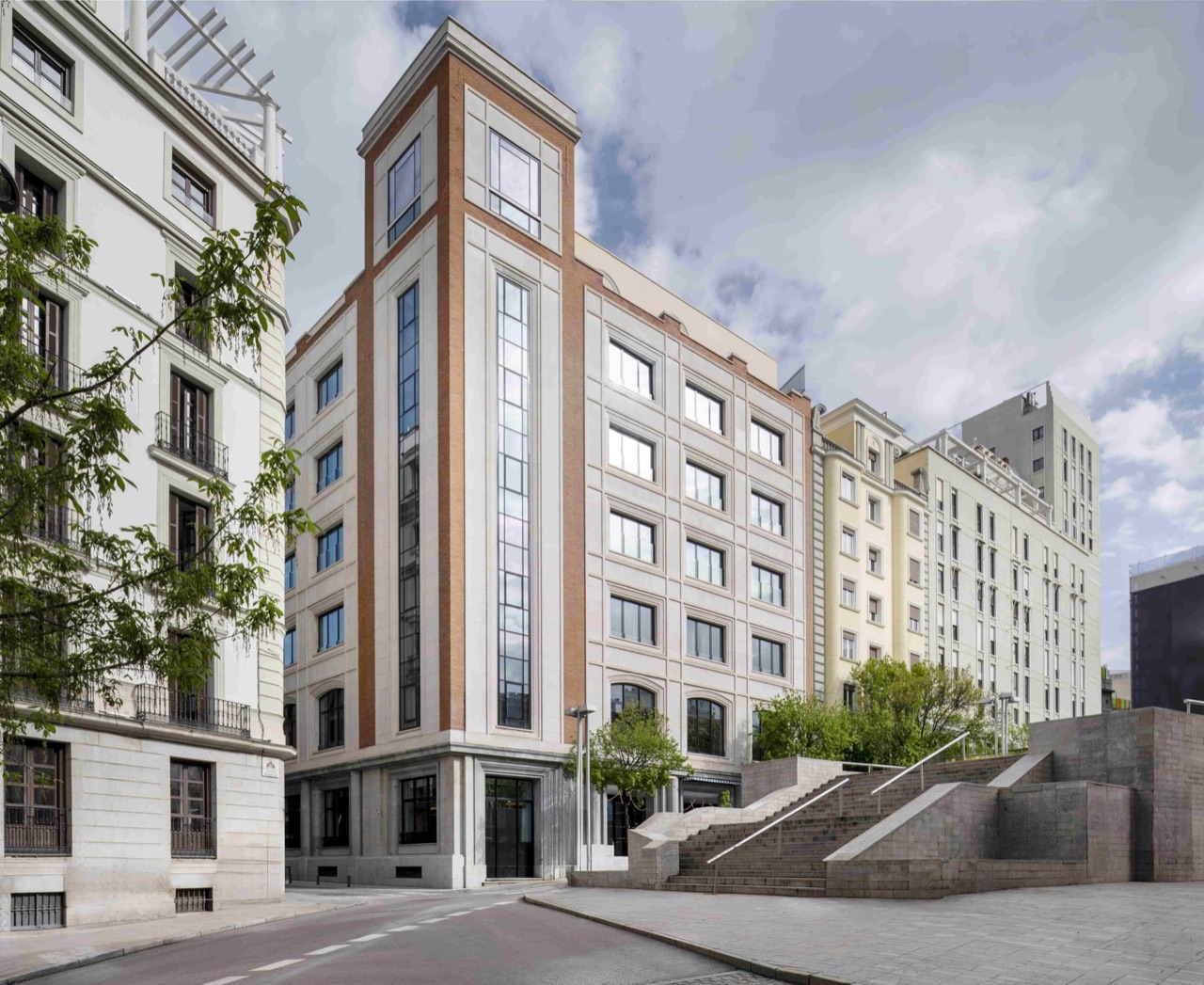 The narrow stone and brick corner facade of Urban Hive Madrid rising above a street, flanked by older apartment buildings.
