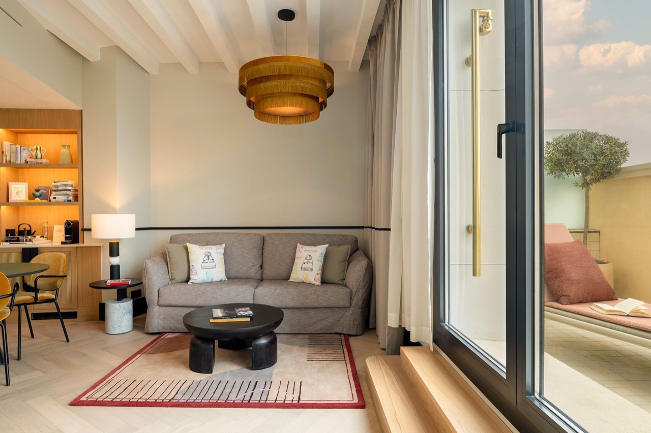 A suite living area with a grey sofa, a built-in bookshelf and coffee station, and glass doors opening onto a private terrace.
