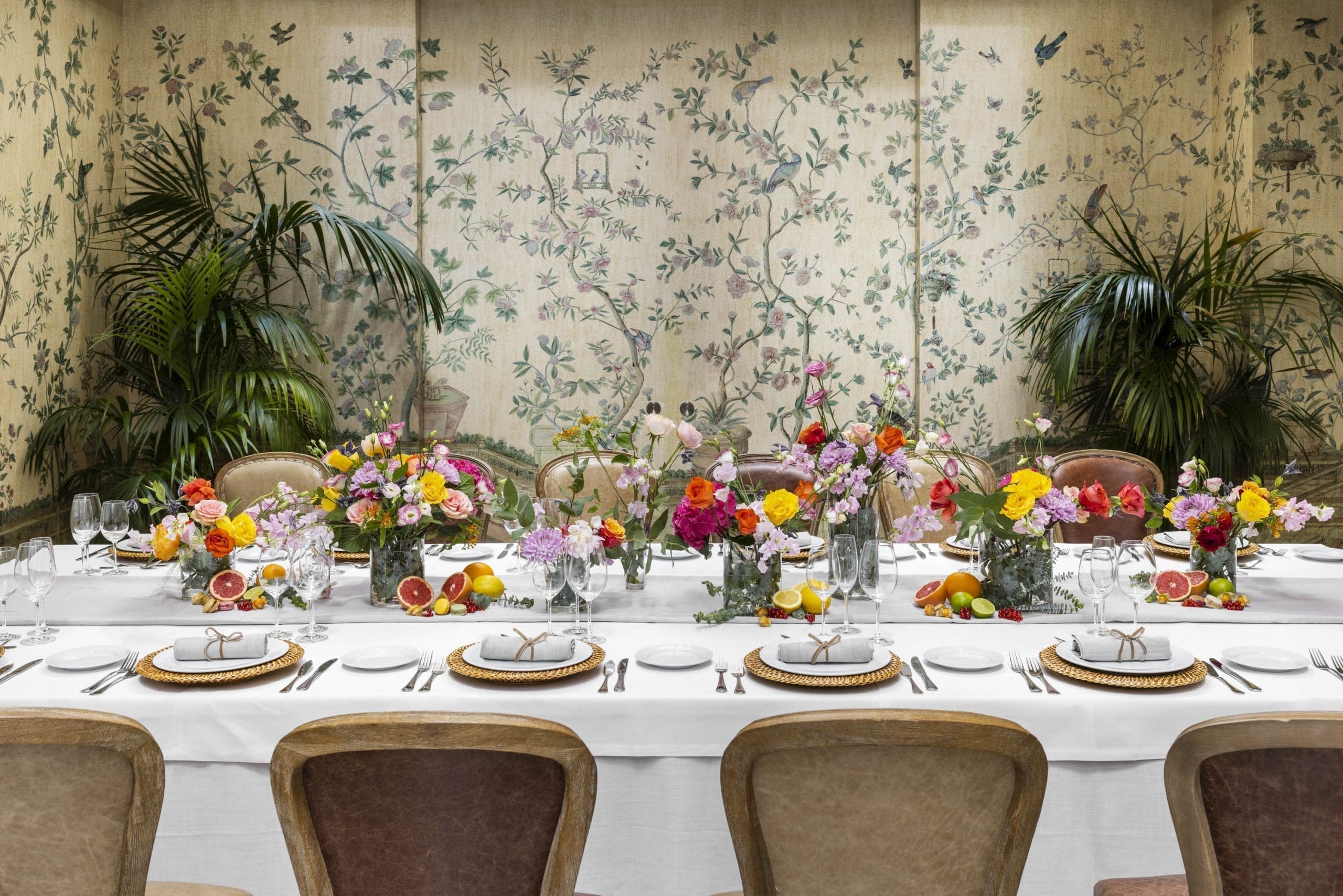 A private dining room with a long white-clothed table set with colourful flower arrangements and citrus fruit, against hand-painted botanical wallpaper and potted palms.