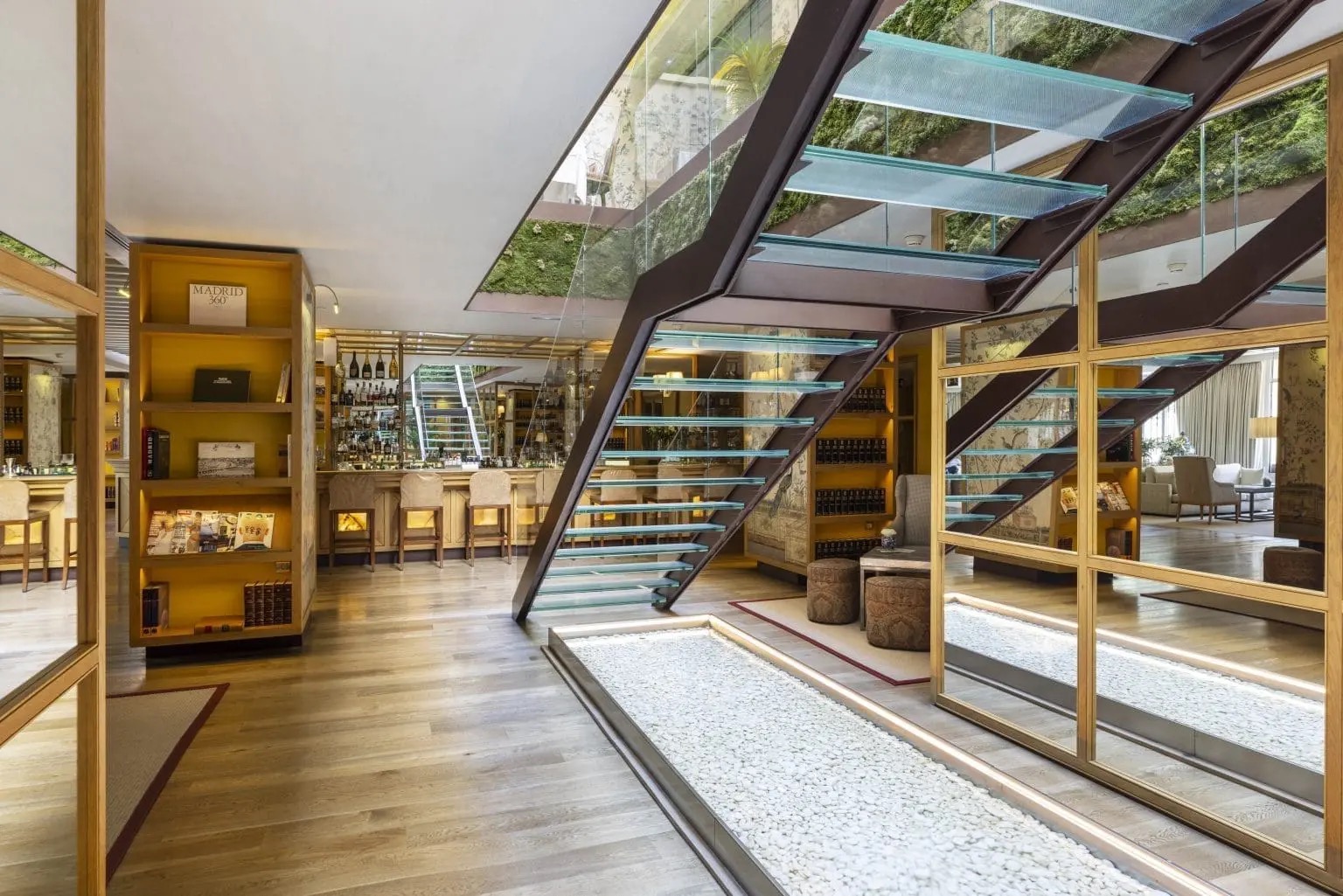 A lobby with a glass-tread staircase beneath a moss-covered wall, a bar counter and stools to one side, and shelves holding books including one titled 'Madrid 360'.