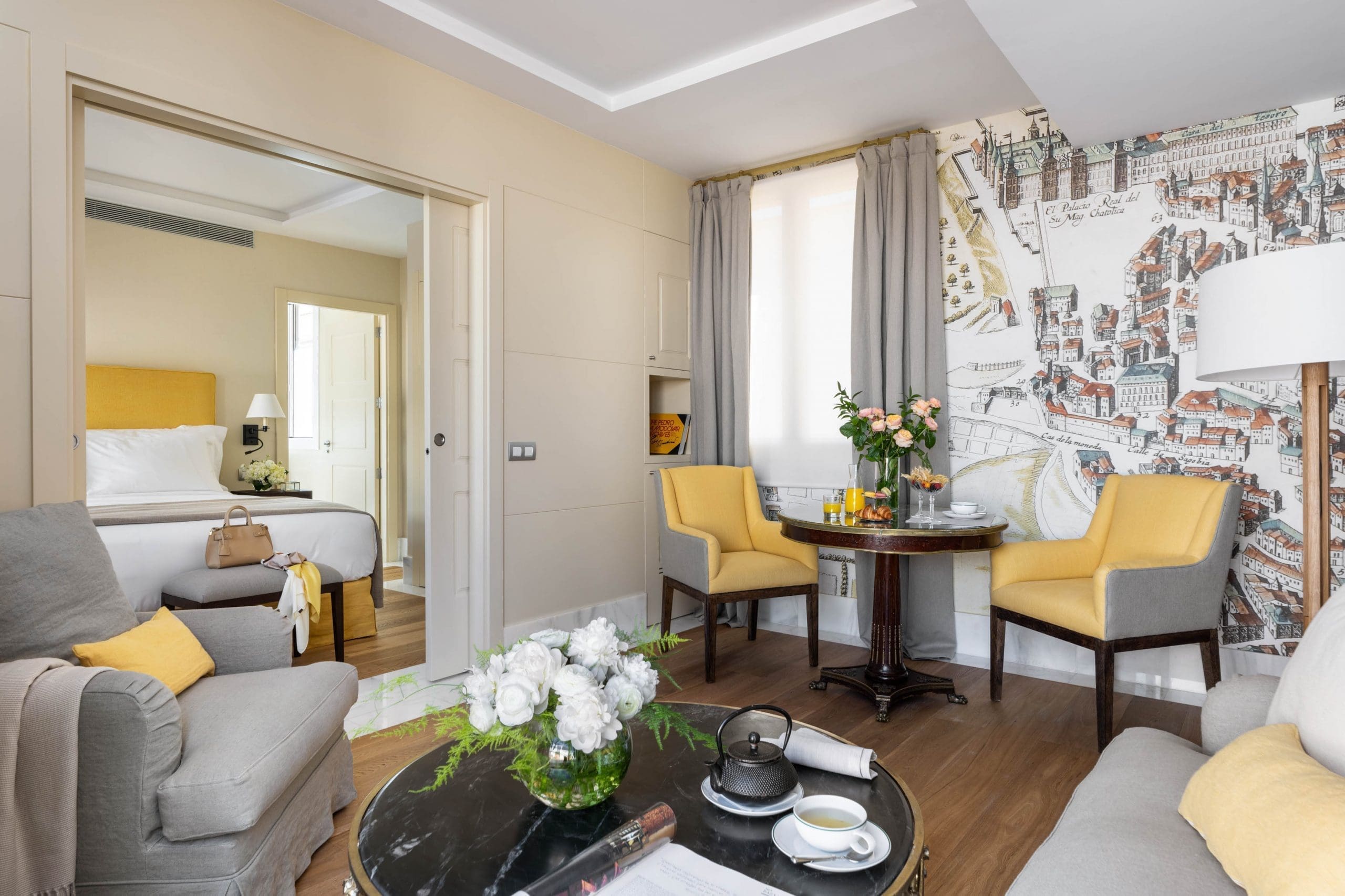 A suite living room with grey sofas, two yellow armchairs at a round breakfast table, an antique map of Madrid covering one wall and a bedroom visible through sliding doors.