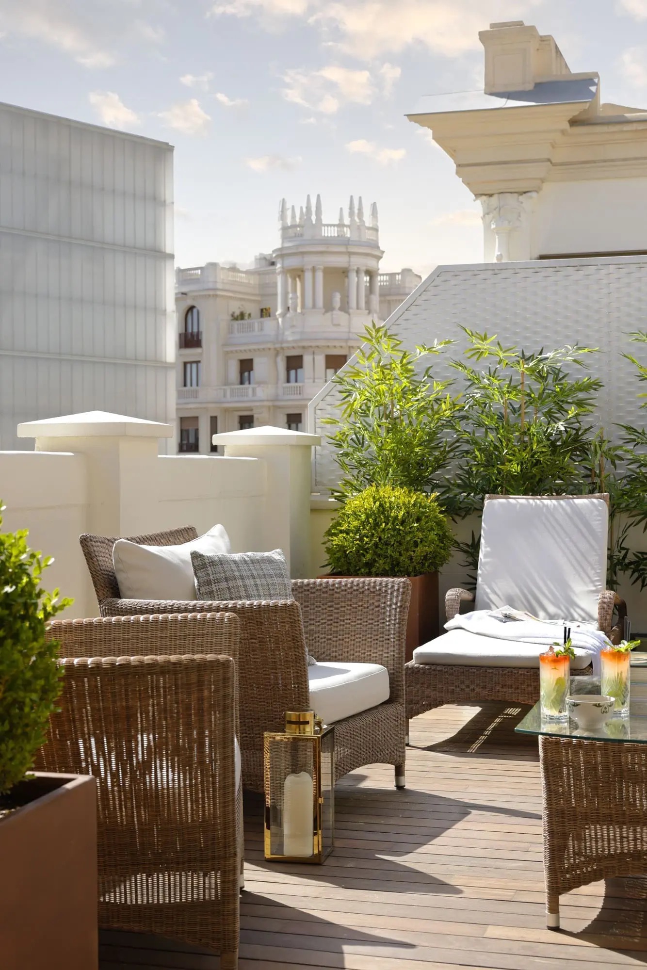 A rooftop terrace with wicker armchairs and a lounger, clipped box and bamboo in planters, two tall cocktails on a glass table and a domed colonnaded building among the rooftops behind.