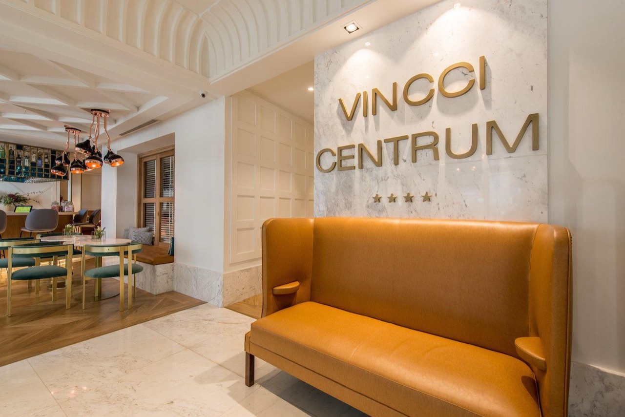 Hotel lobby with a mustard leather bench seat under a marble wall reading VINCCI CENTRUM, with the hall lounge bar and dining tables visible behind it.