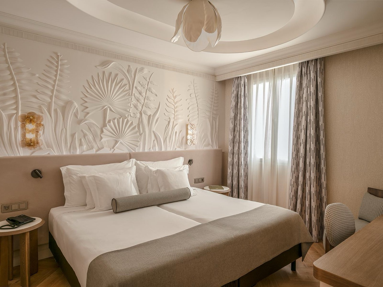 Hotel room with a white-linened bed against a cream relief wall carved with palm and fern leaves, sheer curtains, and a wood writing desk.