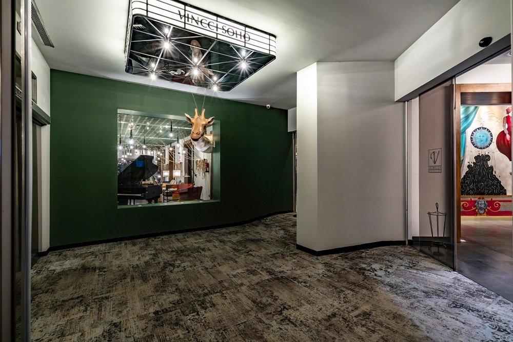 Vincci Soho's lobby corridor with a dark green accent wall, a mounted giraffe-head sculpture and a lit Vincci Soho sign on the ceiling.