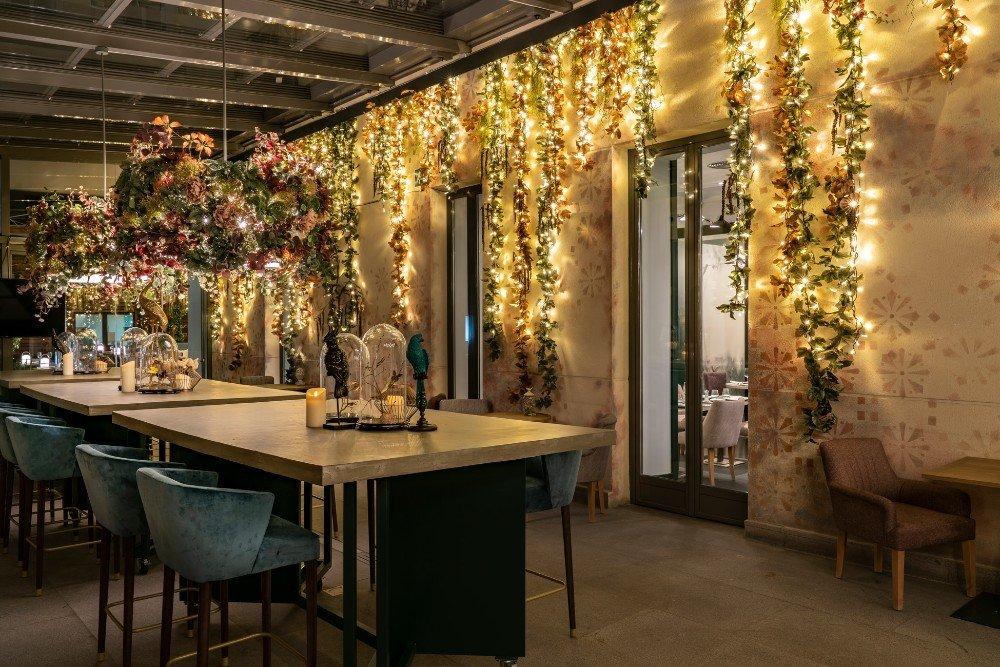 Vincci Soho's restaurant dining area at night, with high-top tables, teal chairs and strings of fairy lights and greenery hanging along the wall.