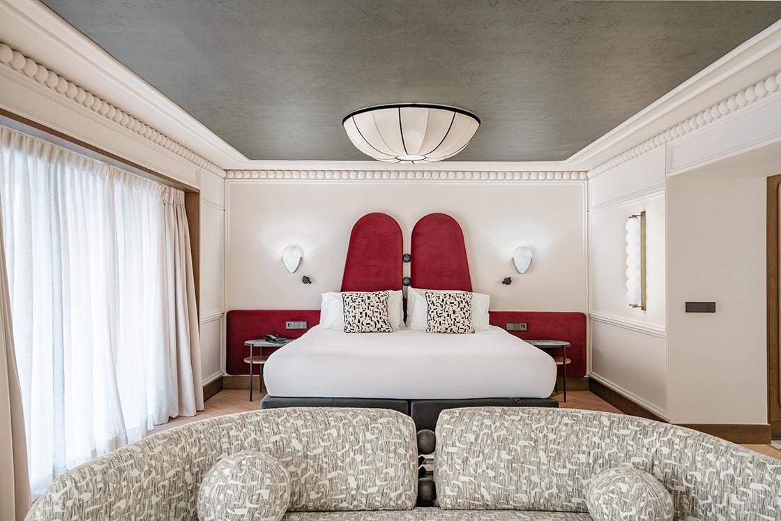 A Suite Deluxe room at Vincci Soho with a dark red padded headboard, patterned armchair and dark ceiling with a round pendant light.
