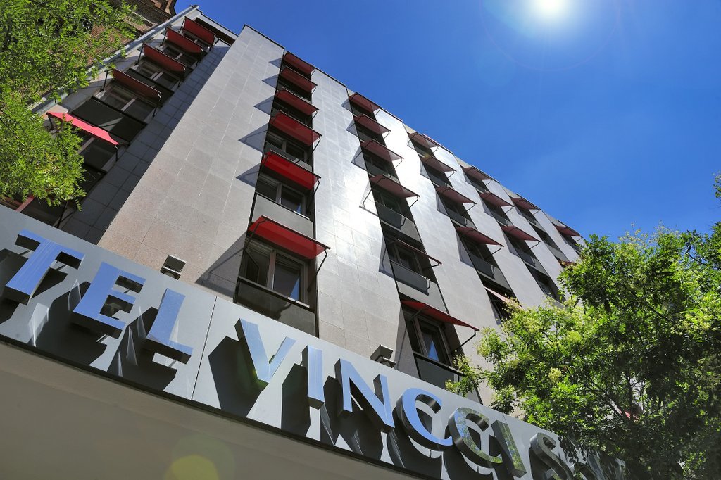 A low-angle view of the Vincci Soma hotel facade, with red-trimmed balconies and the hotel's sign in metal letters against a blue sky.