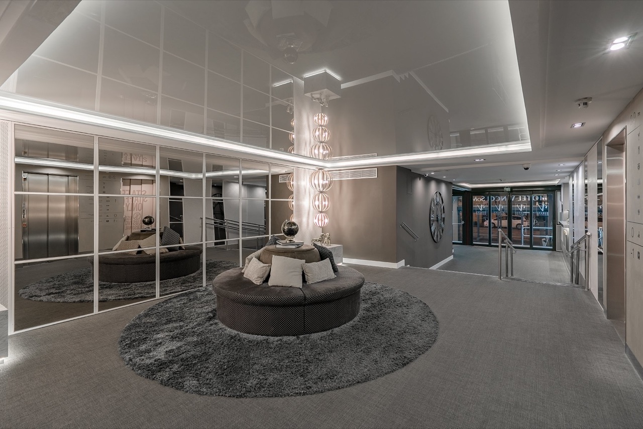 The hotel lobby with a round seating area on a grey rug, mirrored walls reflecting the lift lobby, and the entrance doors visible in the background.