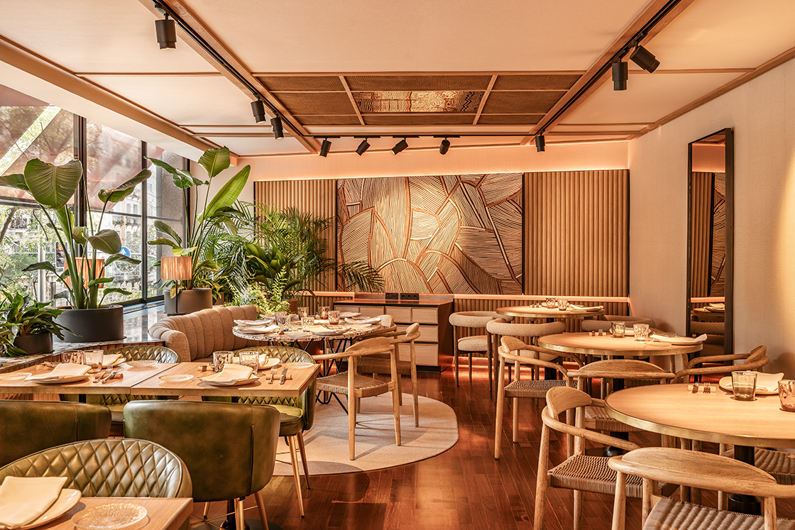 The dining room at SoMa restaurant, with round wood tables, woven chairs and large potted plants in front of floor-to-ceiling windows.