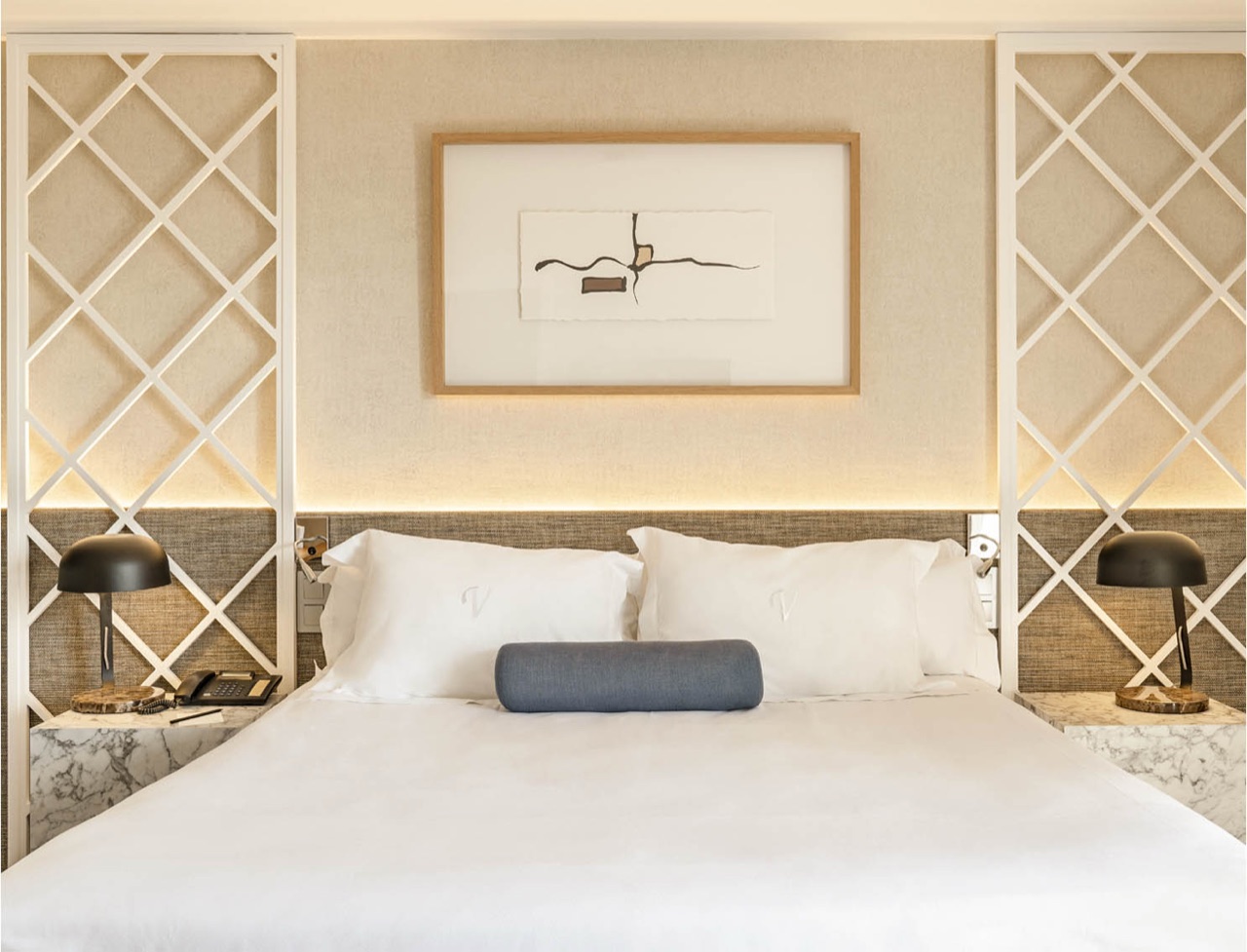 A hotel bed with white linens and a navy bolster pillow embroidered with a V monogram, flanked by two black table lamps on marble nightstands.