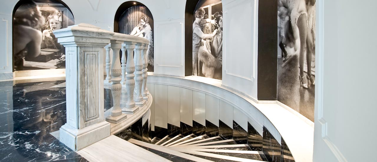 A curved marble staircase and balustrade in the hotel's public area, with large black and white theatre-themed photographs on the walls.