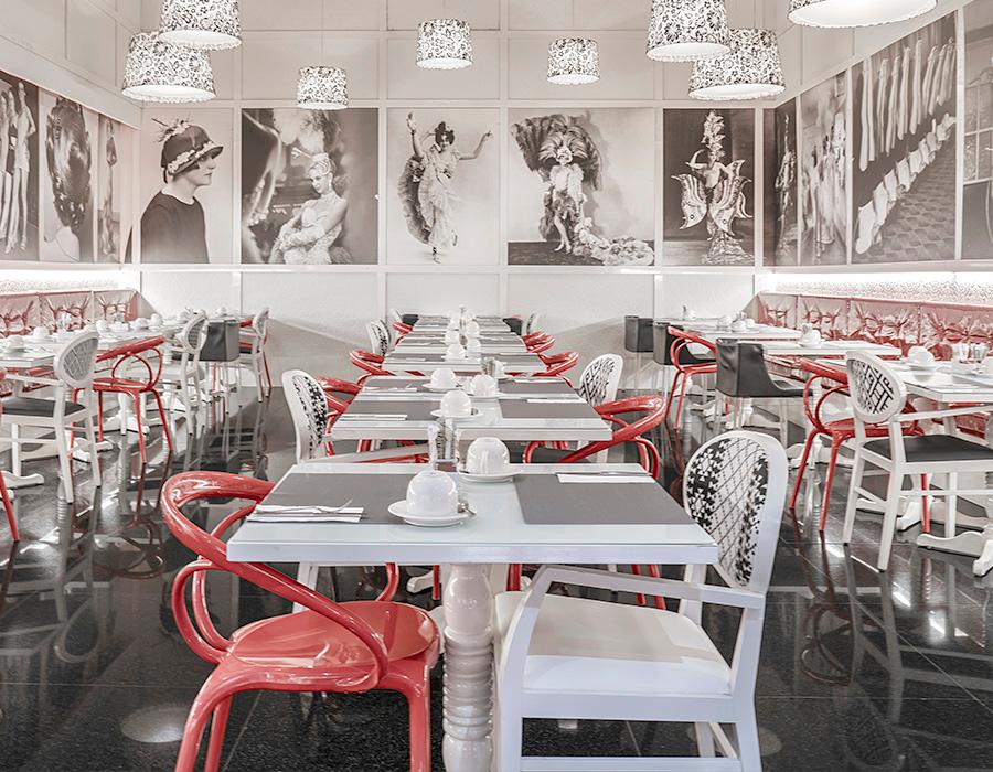 The Le Burlesque dining room with red and white chairs at set tables under patterned pendant lamps, black and white theatre photography on the walls.