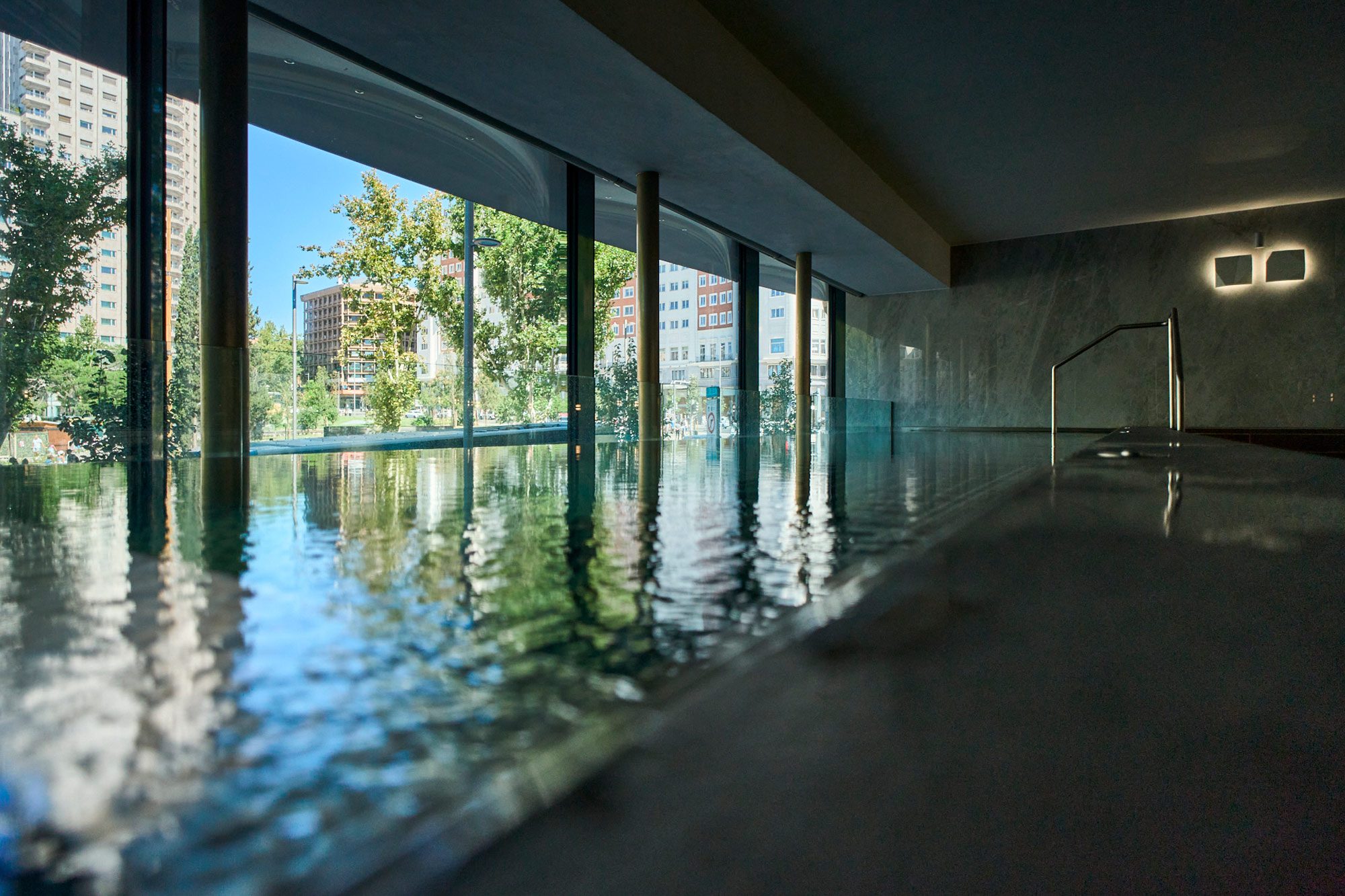 An indoor pool running the length of a full-height glass wall, with marble cladding, a stainless steel handrail and trees and apartment blocks outside.