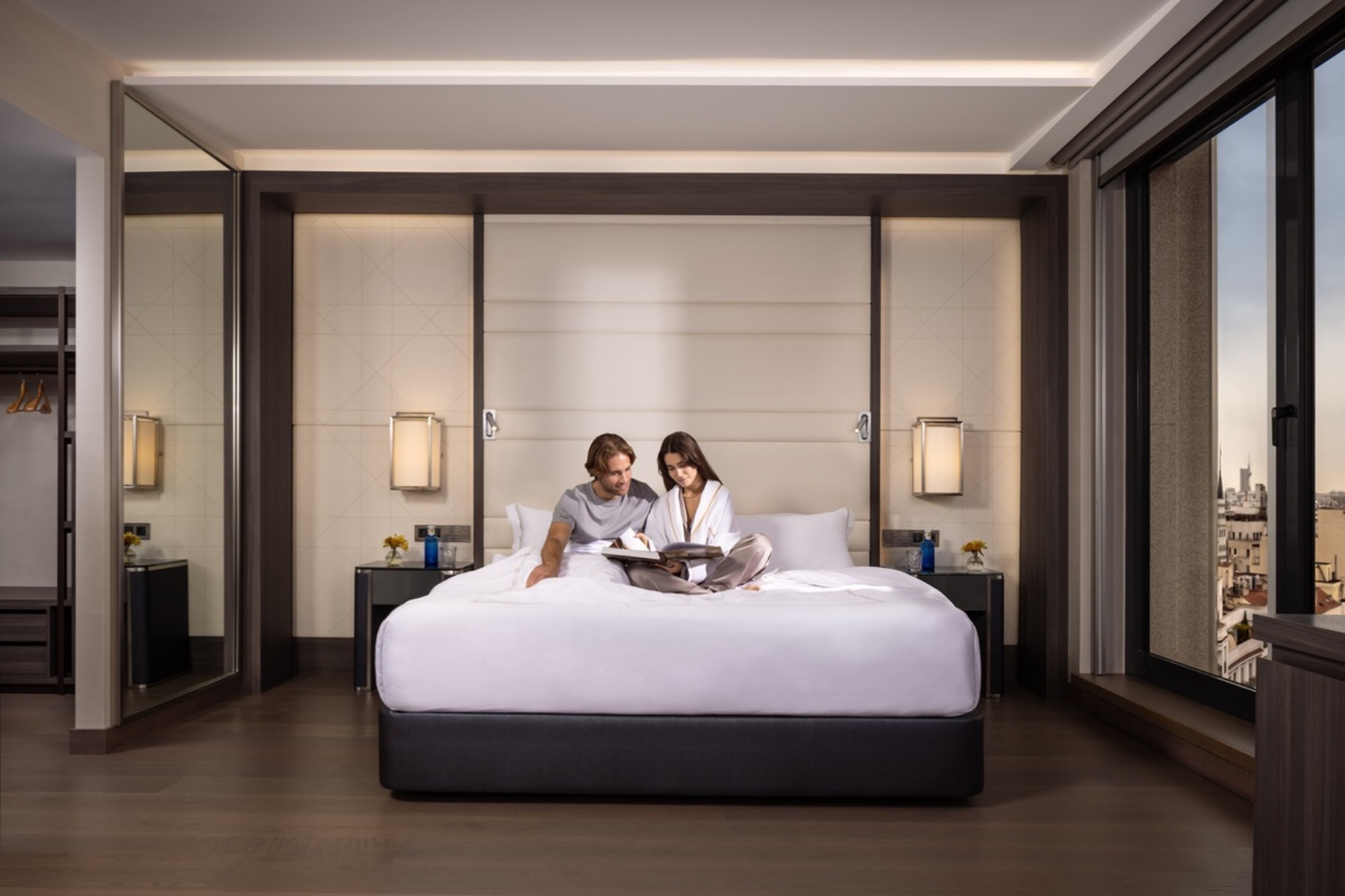A bedroom with a full-width padded headboard wall, matching wall lamps and nightstands, a couple reading on the white bed and the city skyline through the corner window.