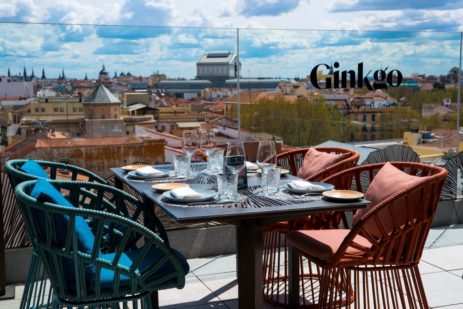 Woven armchairs in blue and terracotta around a set table on a rooftop terrace, with 'Ginkgo Sky Bar' etched on the glass screen and rooftops and spires stretching behind.