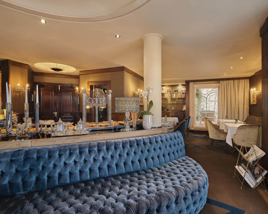 The breakfast room: a tufted blue velvet banquette, a buffet counter with silver teapots and candelabra, and linen-set tables by the window.