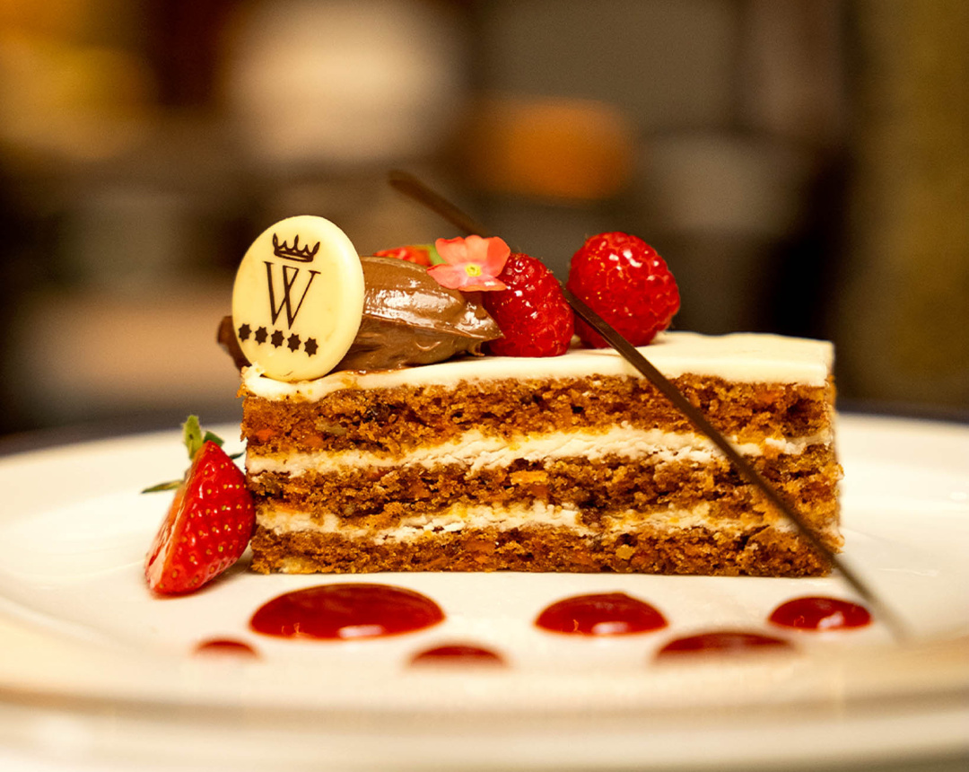 A slice of layered carrot cake plated with strawberries, raspberries, dots of red coulis and a white-chocolate disc bearing the hotel's crowned W emblem.
