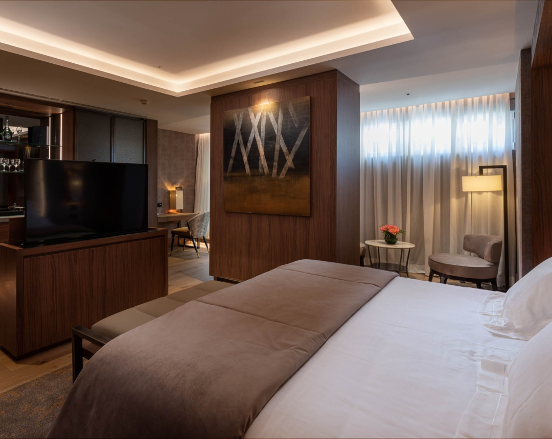 A guest room with a wide bed, a suede headboard wall, a television cabinet with a small bar behind it and a sitting corner lit by a floor lamp.