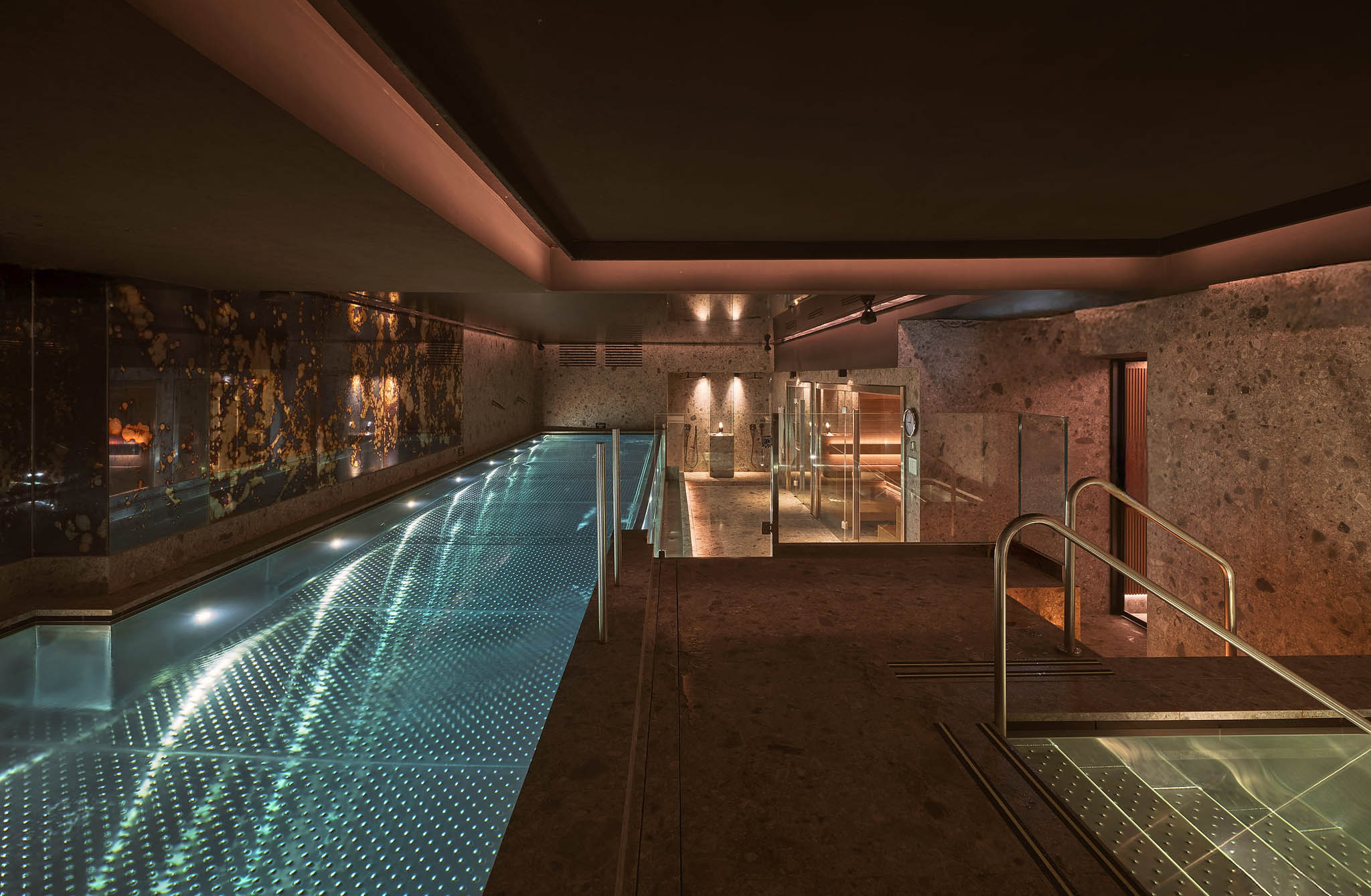 The spa's indoor pool lit from below beside a mottled gold-and-blue mirrored wall, with a glass-walled sauna and shower area at the far end.