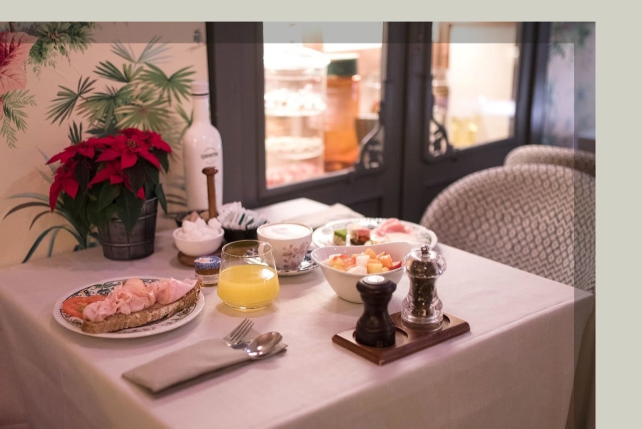 A breakfast table set with a ham and tomato open sandwich, a bowl of fruit, orange juice, coffee, and a potted poinsettia plant.