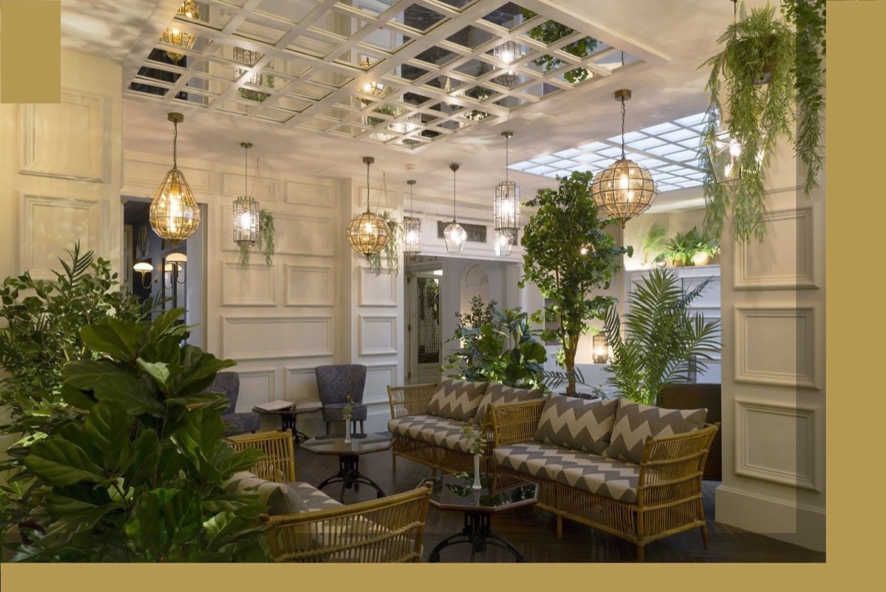 A plant-filled lobby lounge with rattan sofas, a mirrored ceiling grid, hanging lantern lights, and potted greenery throughout.