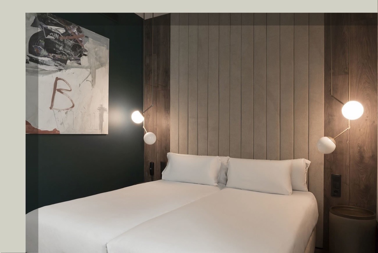 A hotel double room with dark green and wood-panelled walls, twin lamps flanking a white-made bed, and an abstract painting on the wall.