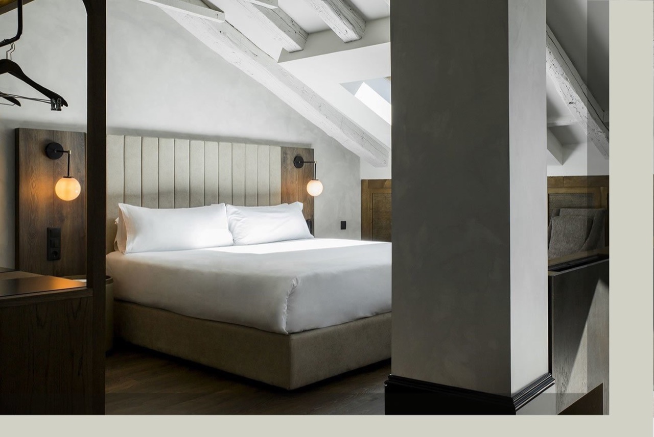 A junior suite under a sloped attic ceiling with exposed white-painted beams, a skylight, and a made bed with wall-mounted globe lamps.