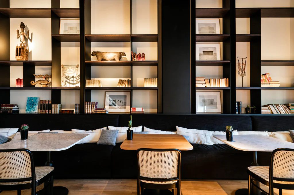 Restaurant at Yurbban Passage with a curated dark wood shelving wall of books and objets, a black banquette, and cane-back chairs at marble tables