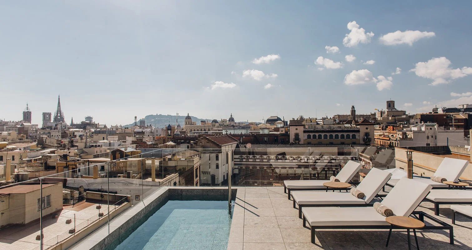 Rooftop pool at Yurbban Passage with glass balustrades, cream sun loungers, and a panoramic view over Barcelona's old town rooftops and spires