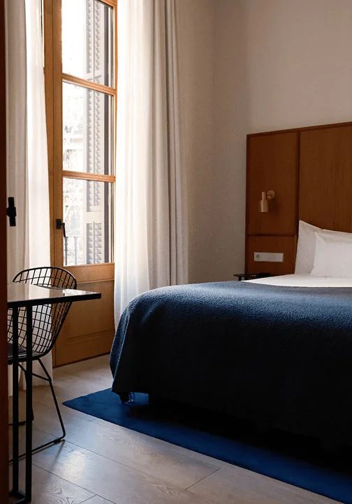 Guest room at Yurbban Passage with a navy bedspread, wood-panelled headboard, and tall wood-framed window opening onto a Barcelona street