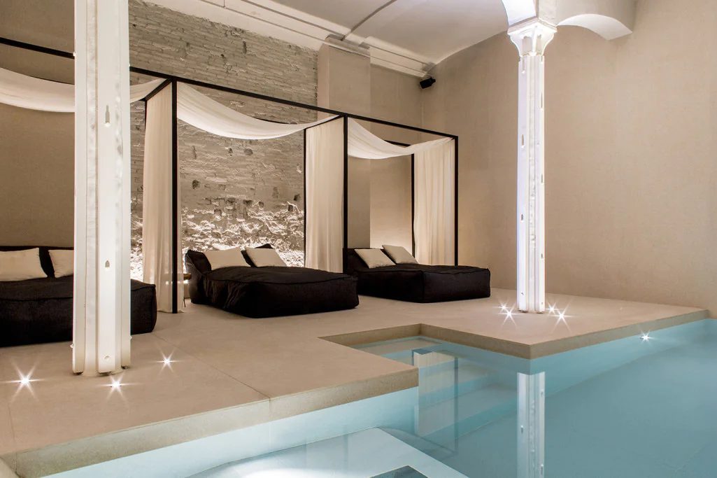 Indoor spa pool at Yurbban Passage with draped canopy daybeds, dark cushions, and an exposed stone wall lit by column lights