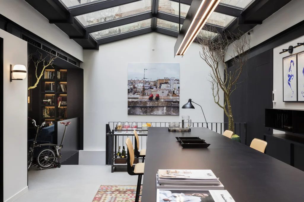 Loft-style interior at Yurbban Ramblas with a black skylight-topped ceiling, built-in bookshelves, a folding bicycle, and a long dark communal table